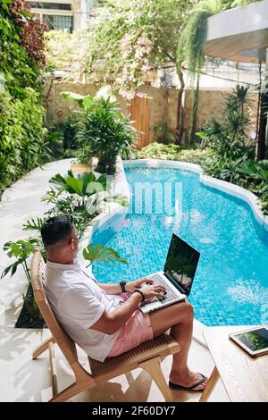 Mann sitzt im Stuhl neben dem Schwimmbad in seinem Hinterhof und arbeitet an Laptop, Freiberufler und Fernarbeit Konzept Stockfoto