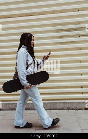 Teenager-Mädchen mit Skateboard mit Smartphone, während zu Fuß auf Fußweg an einer Metallwand Stockfoto