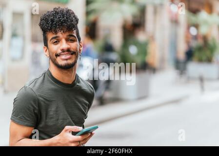 Porträt eines jungen und attraktiven lateinamerikanischen Mannes, der die Kamera anschaut und lächelt. Er hält ein blaues Handy in der Hand. Er ist auf der Straße in Barcelona. Stockfoto