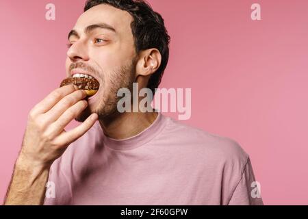 Fröhlich gut aussehend hungrig Kerl essen Donut auf Kamera isoliert über Rosa Hintergrund Stockfoto