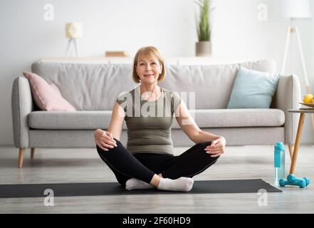 Positive ältere Frau, die in Lotuspose auf Sportmatte sitzt, zu Hause meditiert, Platz kopiert Stockfoto