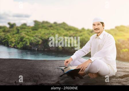 Asiatischer muslimischer Mann sitzt und liest den Koran mit AT Im Freien Stockfoto