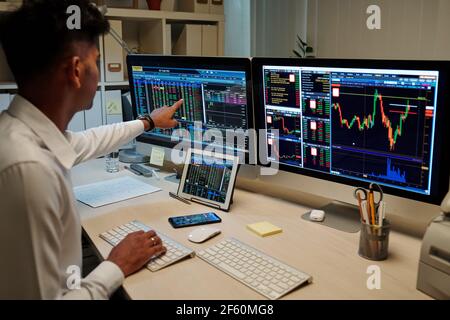 Professioneller junger indischer Händler, der im dunklen Büro arbeitet und zeigt Auf dem Computerbildschirm mit Börseninformationen Stockfoto