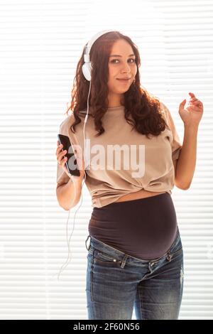 Schöne junge friedliche schwangere Frau hört angenehme klassische Musik mit Smartphone und Kopfhörer. Konzept der positiven Einstellung vor Stockfoto