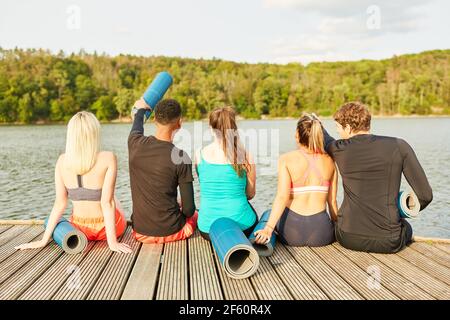 Gruppe von jungen Menschen mit Yoga-Matten sitzen auf einem Holzsteg am Seeufer in der Natur Stockfoto