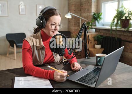 Junge Frau in Kopfhörer sitzen am Tisch arbeiten an Laptop und Aufnahme Podcast sprechen im Mikrofon Stockfoto