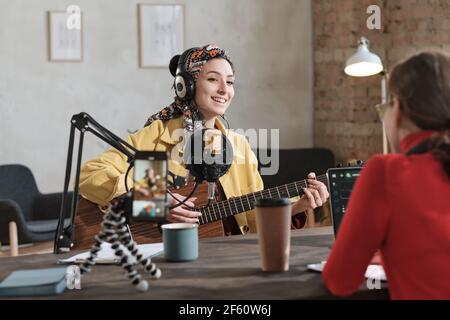 Junge Frau mit Kopfhörern, die Gitarre spielt und ein Lied singt Während der Luft im Radiostudio mit Radio dj sitzt gegenüber Sie Stockfoto