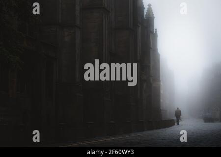 Silhouette eines Mannes, der neben der St Giles' Cathedral in der stimmungsvollen Altstadt von Edinburgh entlang der kopfsteingepflasterten Royal Mile im nebligen Nebel geht. Stockfoto