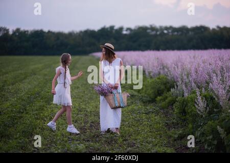 Junge Mutter in weißer Sundress, Strohhut umarmt kleine Tochter. Mädchen gehen mit Frau zwischen blühenden lila Salbei Feld. Mütterliche Fürsorge Liebe. Urlaub mit wi Stockfoto