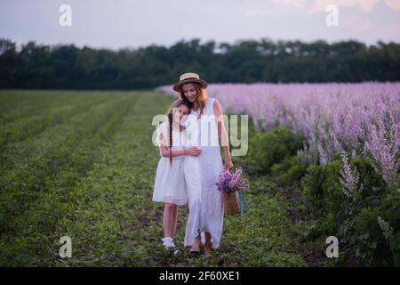 Junge Mutter in weißer Sundress, Strohhut umarmt kleine Tochter. Mädchen gehen mit Frau zwischen blühenden lila Salbei Feld. Mütterliche Fürsorge Liebe. Urlaub mit wi Stockfoto