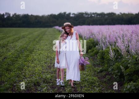 Junge Mutter in weißer Sundress, Strohhut umarmt kleine Tochter. Mädchen gehen mit Frau zwischen blühenden lila Salbei Feld. Mütterliche Fürsorge Liebe. Urlaub mit wi Stockfoto