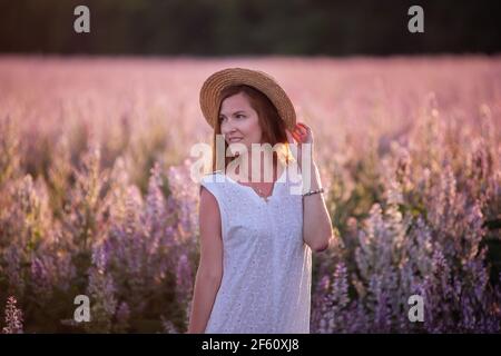 Eine junge Frau in einer weißen Sundress, Strohhut hält einen Weidenkorb mit einem Strauß. Ein Mädchen geht bei Sonnenuntergang durch ein Salbei blühendes rosa Feld. Die Konz Stockfoto