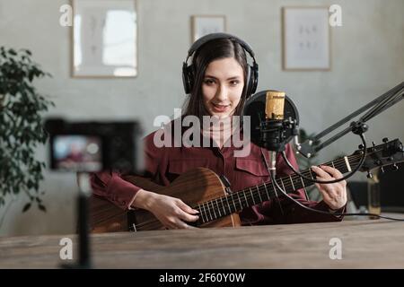 Portrait einer jungen Frau in Kopfhörern, die Gitarre spielt und Aufnahmen Ein Lied im Aufnahmestudio Stockfoto