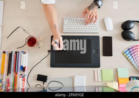 Hände des Grafikdesigners Zeichnung auf Tablet mit digitalem Stift, Smartphone Farbpalette und Tasse Tee herum, Blick von oben Stockfoto