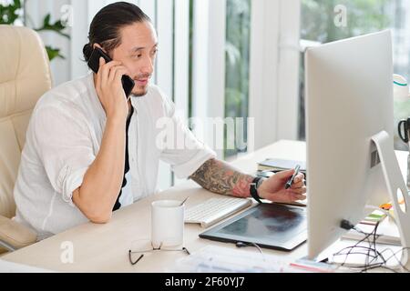 Schöner professioneller Grafikdesigner, der mit dem Kunden telefoniert und Zeichnen mit digitalem Stift Stockfoto