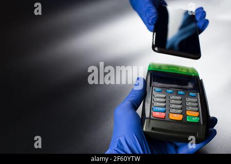 Kontaktloser Bezahlvorgang, Hände in blauen Handschuhen, die kontaktloses Terminal und Smartphone auf anderen Händen halten, auf schwarzem Hintergrund mit Kopierraum. Stockfoto