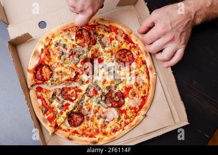 Man Hand schneidet eine appetitliche Pizza in Scheiben, in einer Schachtel, auf einem schwarzen Tisch. Draufsicht. Stockfoto