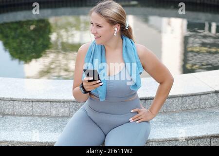 Glücklich ziemlich junge plus size Frau sitzt am Brunnen ruht Nach dem Training im Freien und dem Überprüfen des Smartphones Stockfoto