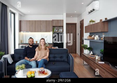Ein glückliches junges Mixed-Race-Paar, das Zeit in ihrer Studiowohnung verbringt, auf dem Sofa sitzt und die Kamera anschaut Stockfoto
