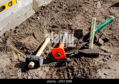 Unschärfe-Hammer, Hammer, Meißel und Bajonett der Schaufel liegen auf dem Boden. Set von Werkzeugen für die Verlegung von Pflastersteinen. Arbeitsprozess. Vorbereitung zum Lay y Stockfoto