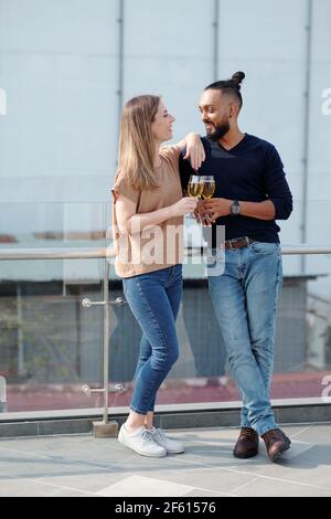 Positives junges Paar, das auf dem Dach steht, Champagner trinkt und sich anschaut Stockfoto