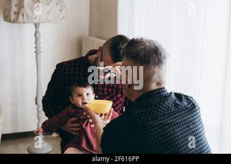 Männlich Gay paar mit angenommen Baby Mädchen zu Hause - Zwei schöne Väter füttern das Baby Mädchen in der Küche - Männliche Babysitter - LGBT Familie zu Hause - Vielfalt Stockfoto