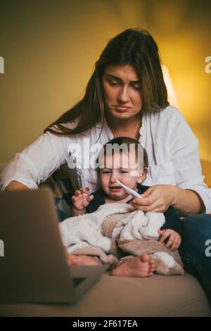 Mutter überprüft die Temperatur der Tochter, während sie online Ratschläge eingibt Laptop zu Hause Stockfoto