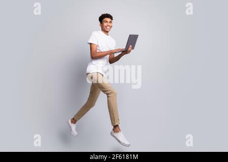 In voller Größe Profil Foto von Brunet optimistisch schönen Kerl springen Halten Laptop tragen weißen T-Shirt Hose isoliert auf grauem Hintergrund Stockfoto