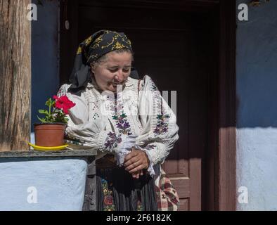Salaj, Transsilvanien, Rumänien-14. Mai 2018: Alte Frau in traditioneller rumänischer Volkstracht auf der Terrasse ihres kleinen charmanten Hauses sitzend Stockfoto