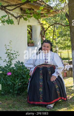 Salaj, Transsilvanien, Rumänien-14. Mai 2018: Alte Frau in traditioneller rumänischer Tracht sitzt im Garten vor ihrem kleinen charmanten Haus Stockfoto