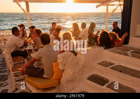 Eine Gruppe von Leuten sitzt in einer Bar im Dorf auf der Insel Mykonos in Griechenland am Wasser bei Sonnenuntergang. Im Hintergrund der helle Himmel und Stockfoto