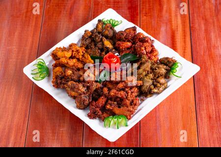 Chicken Pakoda oder Pakora, drei verschiedene Geschmacksrichtungen von Pakora angeordnet in Eine weiße quadratische Platte und garniert mit Tomaten und Gurken Auf einem hölzernen Rücken Stockfoto