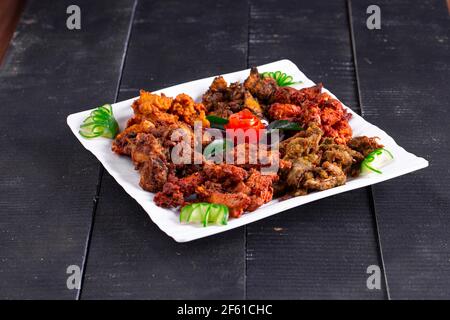 Chicken Pakoda oder Pakora, drei verschiedene Geschmacksrichtungen von Pakora angeordnet in Eine weiße quadratische Platte und garniert mit Tomaten und Gurken Auf einem schwarzen Hintergro Stockfoto