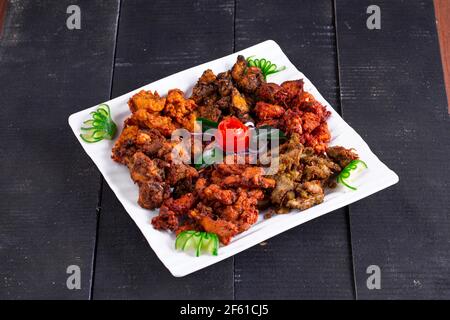 Chicken Pakoda oder Pakora, drei verschiedene Geschmacksrichtungen von Pakora angeordnet in Eine weiße quadratische Platte und garniert mit Tomaten und Gurken Auf einem schwarzen Hintergro Stockfoto