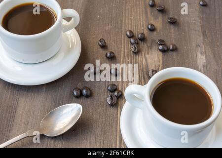 Luftaufnahme von zwei weißen Tassen mit Kaffee und Löffel, auf Holztisch mit Kaffeebohnen, selektiver Fokus, horizontal Stockfoto