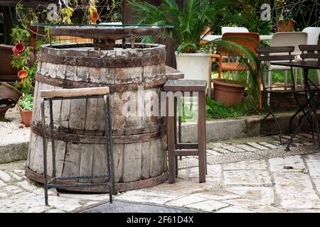 Ein Holz- und Metallweinfass, das als Tisch auf der Holzterrasse des Cafés verwendet wird. Terrasse mit Faßtisch. Ein altes Fass, das als Möbel verwendet wurde. Stockfoto