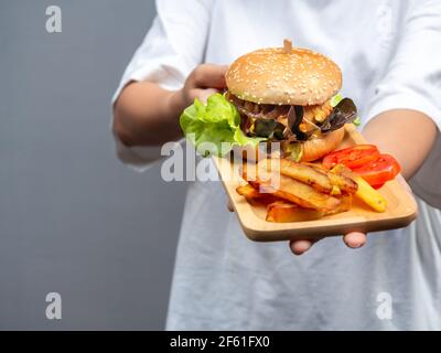 Köstliches, frisch zubereitetes Burger-Set. Frau trägt legeres weißes T-Shirt und zeigt leckeren hausgemachten Hamburger mit pommes Frites und Toma in Scheiben Stockfoto