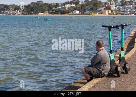 Mann sitzt an der Wand mit zwei Beryl Elektroroller e Scooter escooters in Sandbanks, Poole, Dorset UK im März Stockfoto