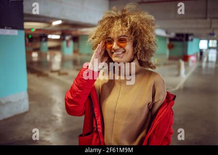 Lächelndes gemischtes Rennen Hip Hop Mädchen in der Jacke stehen in der Garage und Anpassung Sonnenbrille. Stockfoto