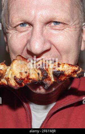 Ein Mann isst ein saftig duftendes Stück Schweinefleisch Kebab auf einem Spieß. Fetthaltige kalorienreiche Lebensmittel. Stockfoto