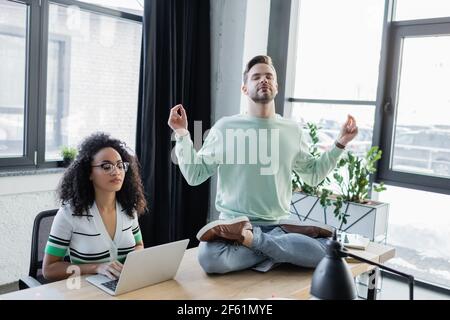 Geschäftsmann sitzt in Yoga-Pose in der Nähe afroamerikanische Geschäftsfrau mit Notebook Stockfoto