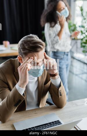 Geschäftsmann, der unter Kopfschmerzen leidet, während afroamerikanischer Kollege darüber spricht Smartphone auf unscharfem Hintergrund Stockfoto