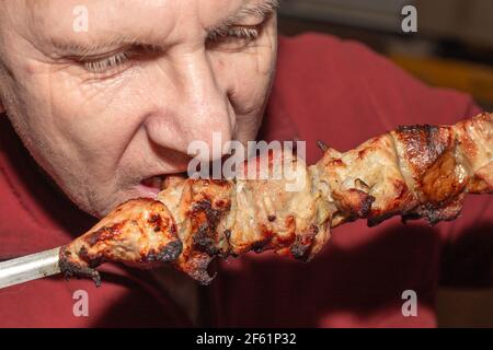 Ein Mann isst ein saftig duftendes Stück Schweinefleisch Kebab auf einem Spieß. Fetthaltige kalorienreiche Lebensmittel. Stockfoto