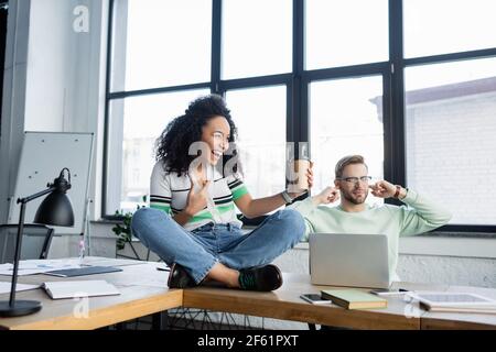 Geschäftsmann bedeckt Ohren neben fröhlichen afroamerikanischen Kollegen mit Kaffee Auf den Bürotisch gehen Stockfoto