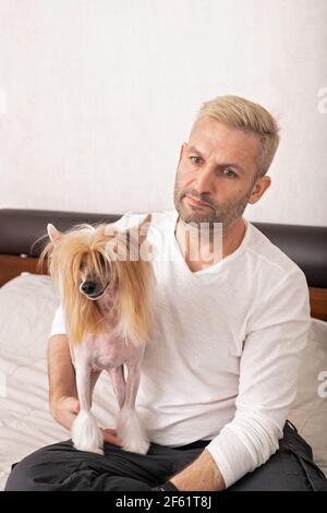 Schöne blonde kaukasischen Mann in einem weißen Hemd hält seine schöne chinesische Haubenhund. Familienhund, Indoor-Aktivität, Lebensstil. Vorderansicht. Stockfoto