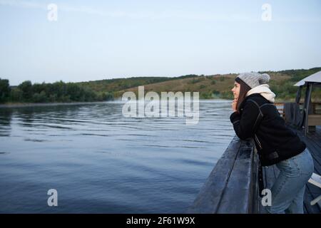 Seitenansicht einer entspannten Frau, die auf dem hölzernen Pier steht und auf die wunderschöne grüne Natur blickt. Hübsche Frau in blauer Jeans, warmer schwarzer Jacke und Strickmütze. Stockfoto