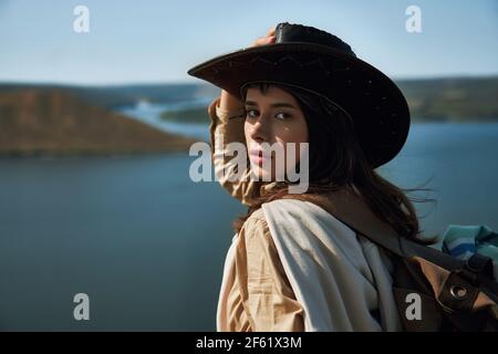 Charmante junge Hündin mit Rucksack auf einem grünen Hügel in der Nähe des breiten Flusses Dniester wandern. Fröhliche Frau im Cowboyhut, die an aktiven Wochenenden im Freien posiert. Stockfoto