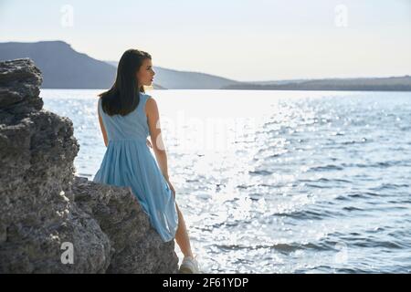 Attraktive dunkelhaarige Frau in stilvollem Kleid sitzt auf Stein in der Bucht von Bakota. Glückliche weibliche verbringen Freizeit auf schöne Natur. Landschaftlich schöner Blick auf den Dniester River. Stockfoto