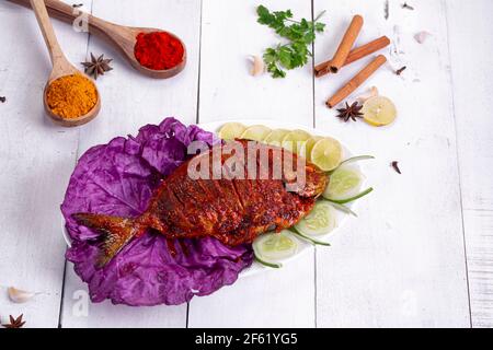 Pomfret braten in einem weißen Teller angeordnet garniert mit lila Kohlblatt und Gurkenscheiben auf weißem strukturierten Hintergrund. Stockfoto