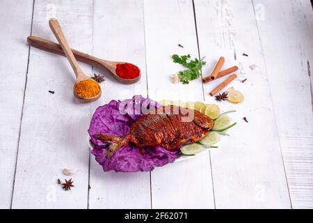 Pomfret braten in einem weißen Teller angeordnet garniert mit lila Kohlblatt und Gurkenscheiben auf weißem strukturierten Hintergrund. Stockfoto